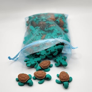 May include: A bag of teal and brown turtle shaped charms. The bag is made of a sheer blue material and is tied with a white string. There are four turtle charms outside of the bag on a white surface.