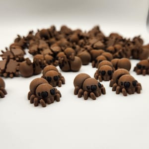 May include: A group of small, brown, 3D printed spiders with black eyes. The spiders are scattered on a white surface.