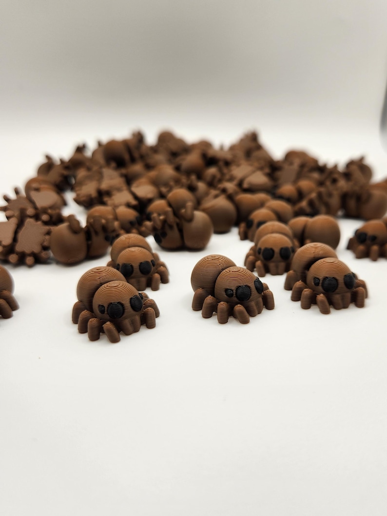 May include: A group of small, brown, 3D printed spiders with black eyes. The spiders are scattered on a white surface.