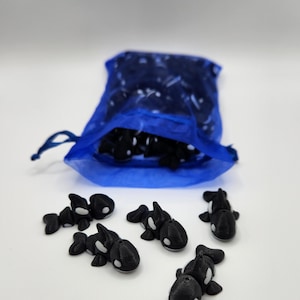 K&ouml;nnte beinhalten: Ein blauer Netzbeutel gef&uuml;llt mit kleinen, schwarz-wei&szlig;en 3D-gedruckten Orca-Figuren. Sechs Orcas liegen verstreut auf einer wei&szlig;en Oberfl&auml;che.