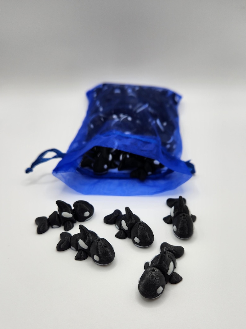 K&ouml;nnte beinhalten: Ein blauer Netzbeutel gef&uuml;llt mit kleinen, schwarz-wei&szlig;en 3D-gedruckten Orca-Figuren. Sechs Orcas liegen verstreut auf einer wei&szlig;en Oberfl&auml;che.