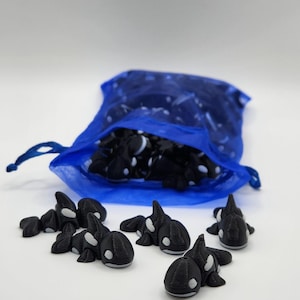 K&ouml;nnte beinhalten: Ein blauer Netzbeutel gef&uuml;llt mit kleinen schwarzen und wei&szlig;en Orca-Figuren. Mehrere Orcas sind auf einer wei&szlig;en Oberfl&auml;che verstreut.