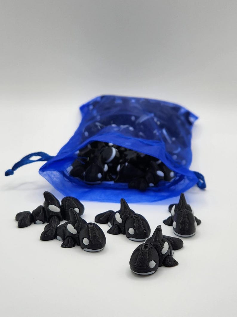K&ouml;nnte beinhalten: Ein blauer Netzbeutel gef&uuml;llt mit kleinen schwarzen und wei&szlig;en Orca-Figuren. Mehrere Orcas sind auf einer wei&szlig;en Oberfl&auml;che verstreut.