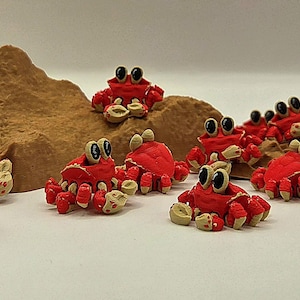 May include: A collection of small, red crab figurines with large, googly eyes and tan claws. The crabs are arranged around brown, textured, miniature rock formations, creating a playful, whimsical scene. The crabs are a bright red color.