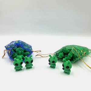 May include: Four small green alien figurines with large black eyes and antennae, two standing in front of a blue star-patterned organza drawstring bag and two standing in front of a green moon and star-patterned organza drawstring bag.