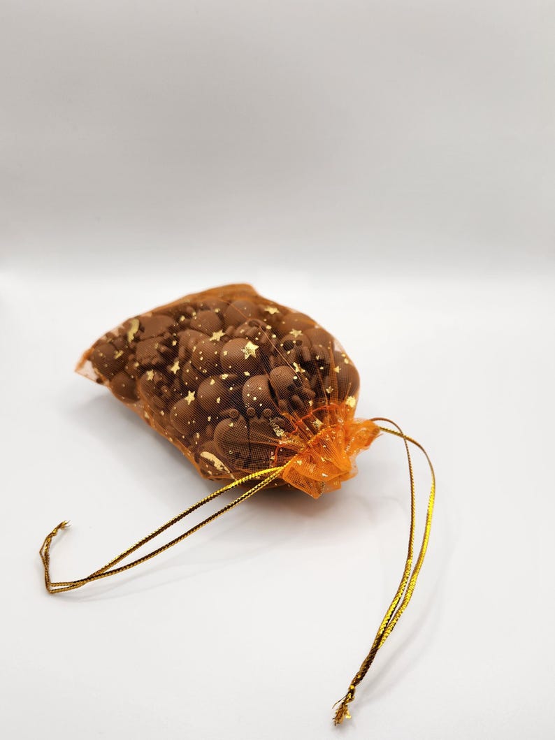 May include: A small orange mesh bag with gold stars printed on it. The bag is filled with small, round, brown chocolate sweets and tied with a gold ribbon.