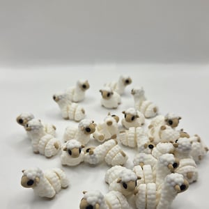 May include: A collection of small, white sheep figurines with tan faces and hooves. The sheep are arranged in a group, with some lying down and others standing. The figurines have a textured, wool-like appearance.