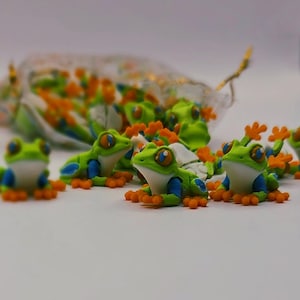 Peut inclure: Plusieurs figurines de grenouilles color&eacute;es. Les grenouilles sont principalement vertes avec des ventres blancs, des d&eacute;tails bleus et des pieds orange. Elles ont de grands yeux d&eacute;taill&eacute;s et sont dispers&eacute;es sur une surface blanche, certaines dans un petit sac.
