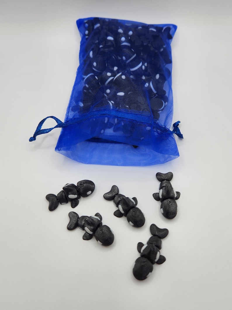 K&ouml;nnte beinhalten: Ein blauer Netzbeutel gef&uuml;llt mit schwarzen und wei&szlig;en Orca-Figuren. F&uuml;nf Orca-Figuren liegen verstreut auf einer wei&szlig;en Oberfl&auml;che.