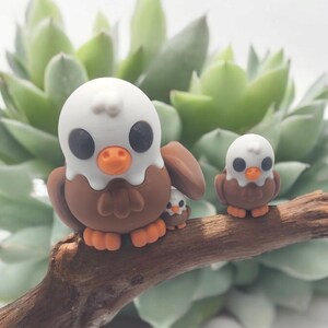 May include: Three brown and white bird figurines with large eyes, perched on a branch. The largest bird has a white head and beak, while the smaller birds have white heads and black beaks.