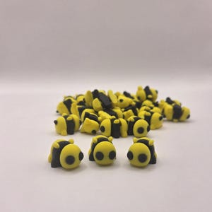 May include: A collection of small, yellow and black bee-shaped erasers. The erasers have a rounded shape with black stripes and small black eyes. The erasers are scattered on a white surface.