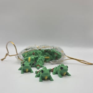May include: A collection of small, green frog figurines with yellow bellies and black eyes. Some frogs are outside a small, clear drawstring bag. The frogs have a textured appearance and are arranged on a white surface.