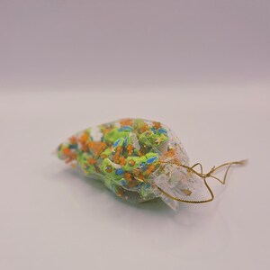Peut inclure: Un petit sac transparent rempli de breloques en forme de grenouilles vertes, oranges et bleues, avec des d&eacute;tails dor&eacute;s. Le sac est nou&eacute; avec un cordon dor&eacute;. Les breloques sont dans un sac transparent sur une surface blanche.