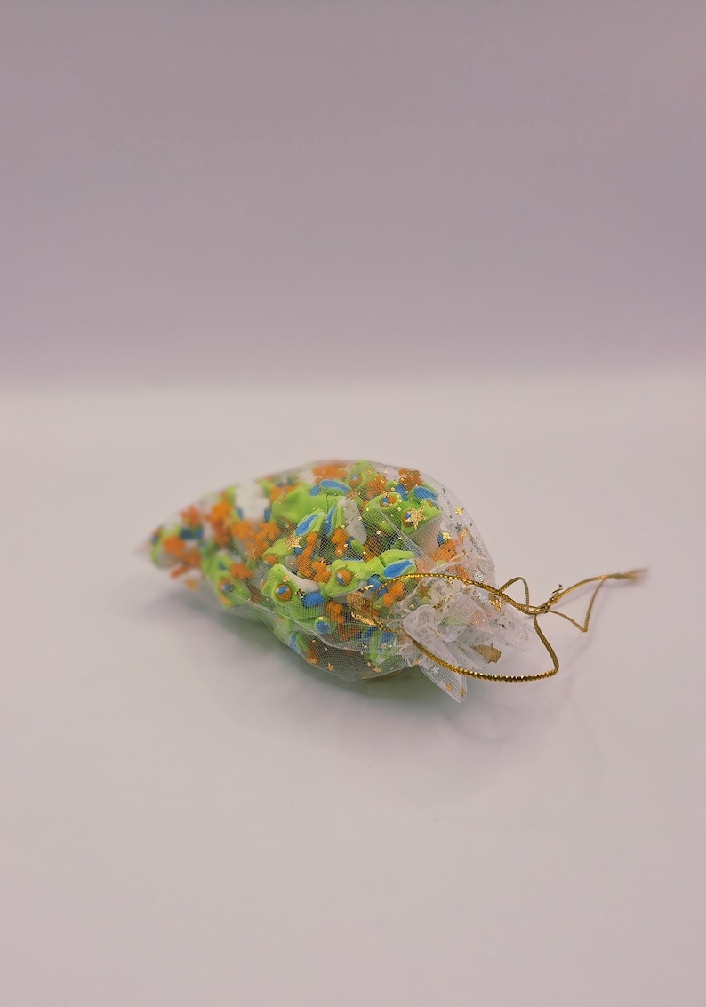 Peut inclure: Un petit sac transparent rempli de breloques en forme de grenouilles vertes, oranges et bleues, avec des d&eacute;tails dor&eacute;s. Le sac est nou&eacute; avec un cordon dor&eacute;. Les breloques sont dans un sac transparent sur une surface blanche.