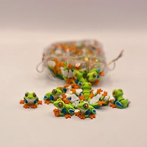 May include: A collection of small, green frog figurines with orange feet and blue accents. The frogs have large, expressive eyes and are scattered around a clear container filled with colorful sprinkles. The figurines are on a white surface.