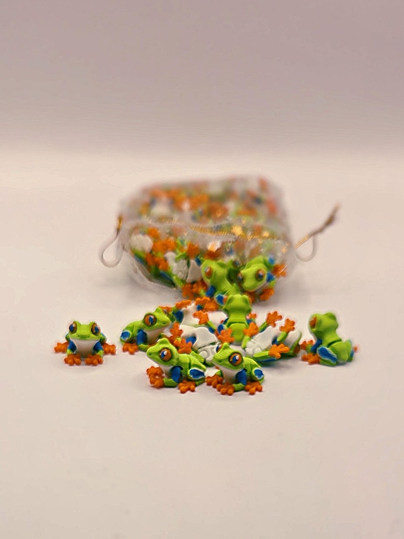 Peut inclure: Petites figurines de grenouilles vertes avec des pieds orange et des d&eacute;tails bleus. Les grenouilles ont de grands yeux expressifs et sont dispers&eacute;es autour d'un r&eacute;cipient transparent rempli de paillettes color&eacute;es. Sur une surface blanche.