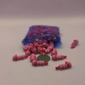 May include: A collection of pink axolotl-shaped charms with red accents, scattered around a blue mesh bag with gold trim. The charms are next to a coin, suggesting size. These miniature axolotls are likely for crafting or jewelry making.