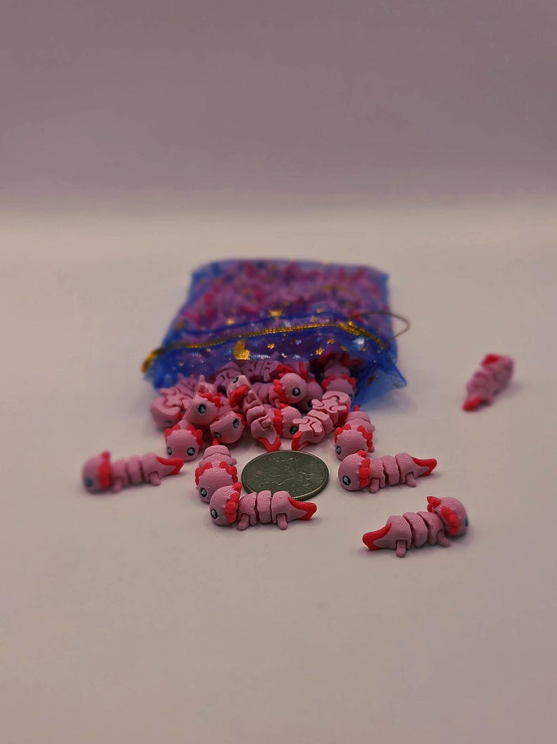 May include: A collection of pink axolotl-shaped charms with red accents, scattered around a blue mesh bag with gold trim. The charms are next to a coin, suggesting size. These miniature axolotls are likely for crafting or jewelry making.