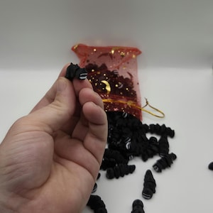 May include: A hand holding a small, black, worm-shaped bead with a white and black mouth design. A red mesh bag with gold stars and more black beads are in the background. The beads are scattered on a white surface.