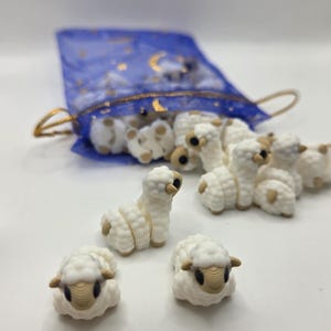May include: A collection of small, white sheep figurines with tan faces and ears. Some sheep are upright, others are lying down. They are spilling out of a blue mesh bag with gold accents.