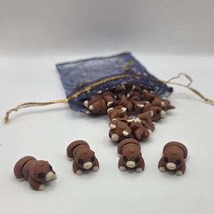 May include: A collection of small, brown bear-shaped erasers spilling from a blue mesh bag with gold accents. Four erasers are in the foreground, each with a white snout and paws. The erasers are on a white surface.