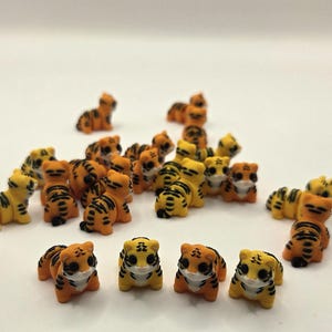 May include: A collection of small, orange and yellow tiger figurines with black stripes. The figurines have white faces and are scattered across a white surface. The tigers are in various poses, some standing and others lying down.