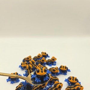 May include: A collection of small, detailed frog figurines. The frogs have a vibrant color scheme of orange and black stripes with blue feet. A gold spoon holds one of the frogs, suggesting a miniature scale.