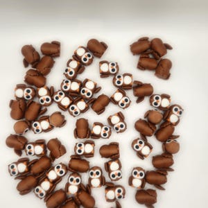 May include: A large group of brown owl-shaped charms with white eyes and orange beaks. The charms are scattered on a white surface.