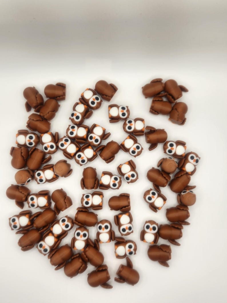 May include: A large group of brown owl-shaped charms with white eyes and orange beaks. The charms are scattered on a white surface.