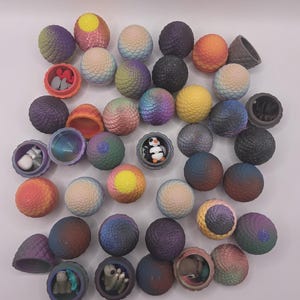 May include: A collection of colorful, spherical toys, some opened to reveal small figurines. The spheres have textured surfaces and are in shades of purple, blue, yellow, and orange. Some toys are open, displaying miniature figures inside.