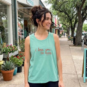 May include: A light teal tank top with the text "i love you so much. Austin, TX" in red script. The tank top is worn by a person standing on a sidewalk lined with trees and shops.