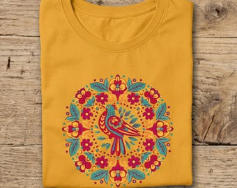 May include: Mustard yellow t-shirt featuring a colorful bird and floral design. The bird is teal, red, and yellow, surrounded by a circular pattern of red flowers, teal leaves, and yellow accents. The shirt is folded on a wooden surface.
