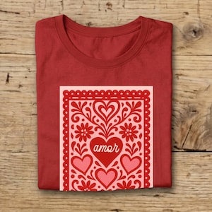 May include: Red t-shirt with a decorative design featuring hearts, flowers, and the word "amor" in a heart-shaped centerpiece. The design is in shades of red and pink, set against a light pink background.
