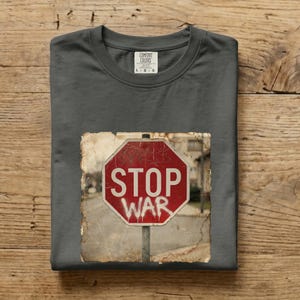 Op de afbeelding: Donkergrijs T-shirt met een versleten afbeelding van een rood stopbord met de witte tekst "STOP WAR". Het shirt is opgevouwen en heeft een label in het midden bovenaan.