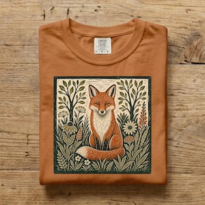 Peut inclure: Un t-shirt orange brûlé avec une impression graphique carrée représentant un renard assis dans un champ d'arbres et de fleurs stylisés. Le motif utilise des tons vert foncé et crème. Le t-shirt porte une étiquette Comfort Colors au col.