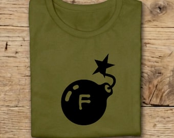 Pode incluir: Camiseta verde-oliva com um gráfico preto de uma bomba, uma estrela e a letra "F". A camiseta está dobrada sobre uma superfície de madeira.