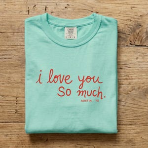 Może przedstawiać: Jasnozielony t-shirt z krótkim rękawem z czerwonym napisem kursywą i love you So much. oraz AUSTIN . TX wydrukowanym poniżej mniejszymi czerwonymi wielkimi literami. Koszulka posiada metkę marki Comfort Colors przy wewnętrznym dekolcie.