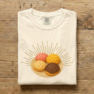 May include: Cream-colored t-shirt featuring a plate of four colorful conchas, a traditional Mexican sweet bread. The conchas are pink, yellow, beige, and brown, with a sunburst design behind them. The shirt has the text "CONENT ENDSS" and "1-06".