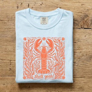 May include: Light blue t-shirt with a screen-printed design featuring an orange lobster surrounded by seaweed and seashells. The text "shell yeah!" is below the lobster. The shirt is folded on a wooden surface.