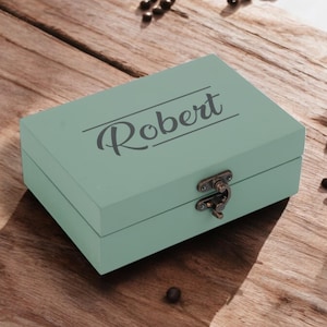 May include: A light green wooden box with a hinged lid and a metal clasp. The box is engraved with the name "Robert".