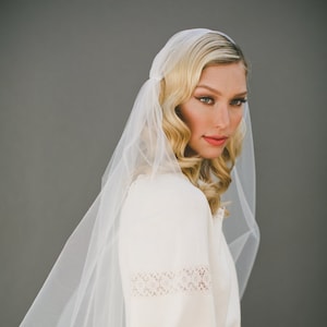 Könnte beinhalten: Eine Braut in einem weißen Brautkleid mit Spitzendetails und einem weißen Schleier. Der Schleier ist über ihren Kopf und ihre Schultern gelegt, und sie schaut zur Seite.