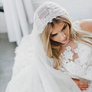 May include: A bride wearing a white lace wedding dress with a white veil and a lace headpiece.