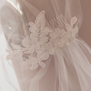 May include: A white bridal veil with a delicate lace applique featuring floral designs. The veil is attached to a comb for easy placement in hair.