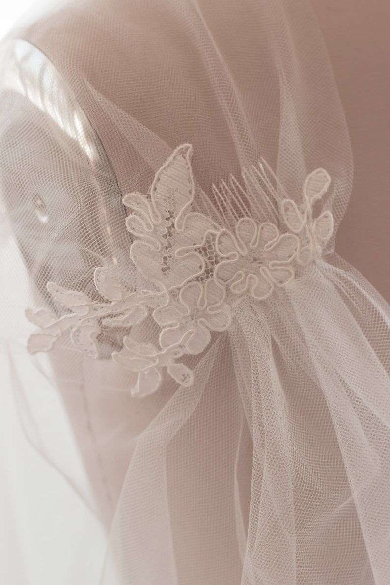 May include: A white bridal veil with a delicate lace applique featuring floral designs. The veil is attached to a comb for easy placement in hair.