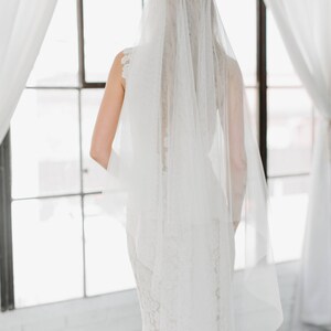 May include: A bride wearing a white wedding dress with a long veil. The bride is standing in front of a window with white curtains.