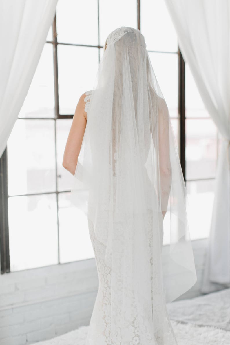 May include: A bride wearing a white wedding dress with a long veil. The bride is standing in front of a window with white curtains.