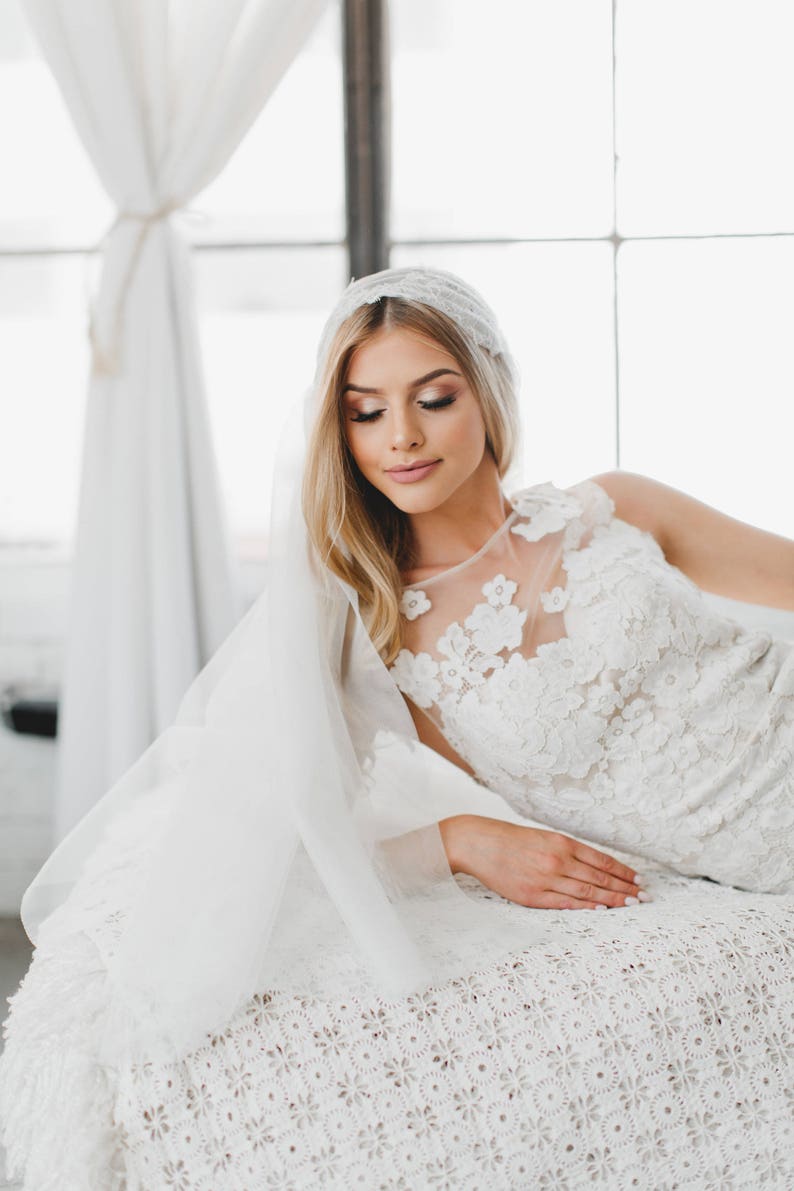 May include: A bride wearing a white lace wedding dress with a veil and a floral headpiece. She is sitting on a white crocheted surface.