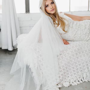 May include: A bride wearing a white lace wedding dress and a white veil with a lace trim. She is lying on a white crocheted blanket.
