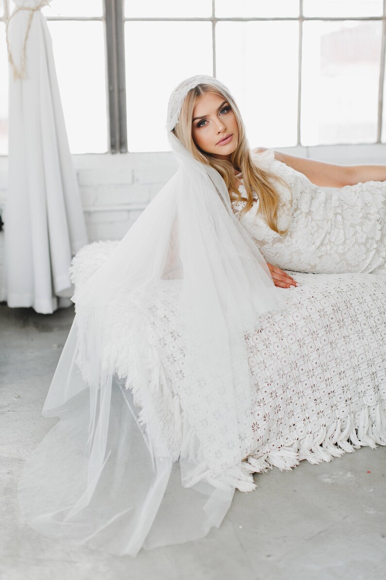 May include: A bride wearing a white lace wedding dress and a white veil with a lace trim. She is lying on a white crocheted blanket.