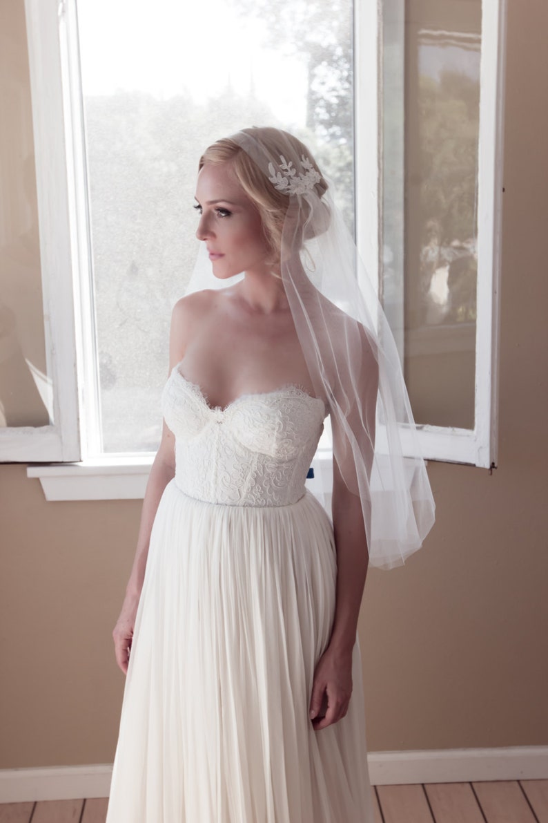 May include: A bride wearing a white strapless wedding dress with a lace bodice and a flowing skirt. She is wearing a white veil with a floral design and has her hair pulled back.